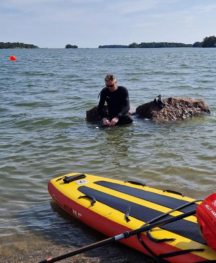 Minä märkäpuvussa istumassa puoliksi veden alla olevan kiven päällä pää painuksissa. Etualalla pelastuskäyttöön tarkoitettu sup-lauta.
