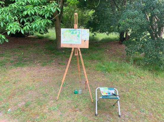Standing easel and watercolour painting setup positioned in front of leafy trees in the Cambridge Botanic Gardens, with paints and materials arranged on a small folding stool.