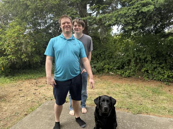 Picture of ACB Membership Services Administrative Assistant Michael Babcock (front) with his son Nicholas (back) standing behind him. To Michael’s left is retired guide dog Titan, a black Labrador retriever. The group stands in a driveway with trees in the background.