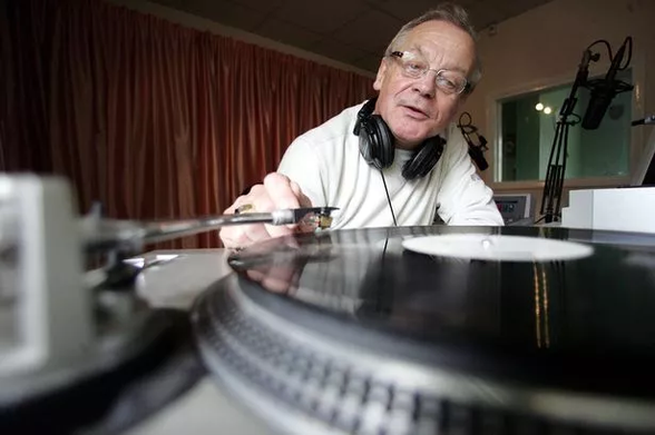 A middle-aged man wearing glasses puts the needle on a vinyl record, which is in the foreground.