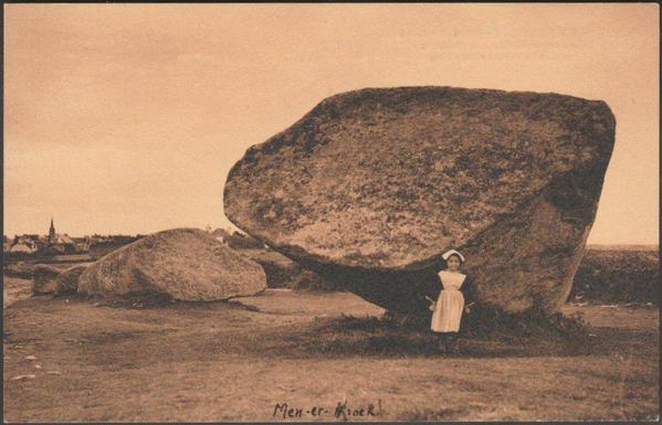 A printed vintage postcard of the Grand Menhir Brisé at Locmariaquer in Brittany. The menhir is, or was, about 23 metres or 75 feet in length and weighs approximately 350 tons. It fell at an unknown date and lies broken into four pieces. It is reputed to be the largest menhir in the world. A young girl in traditional Breton costume stands in front of the closest piece. The town can be seen in the distance. The stone sits within a large complex of prehistoric monuments: the Er Grah tumulus and La Table des Marchands are very close by, and Gavrinis is fairly close, and it may possibly have originally been one of 19 menhirs forming a stone row, the holes for which can still be seen.