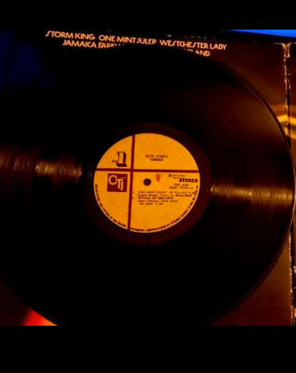  The photograph warmed up with a 81A filter, shows a vinyl record with a yellow label in the center. The label features the text "Bob James Three" and "CTI" logos, indicating the artist and record label. The record is identified as stereo and has a catalog number of CTI 4042. The track titles visible on the label include "Storm King," "One Mint Julep," "Westchester Lady," and "Jamaica Farewell." The record is partially inside its sleeve, which is visible at the top of the image. The background is dark, with a warm, orange glow illuminating the record and label, creating a vintage aesthetic.

 Ovis2-8B

🌱 Energy used: 0.166 Wh