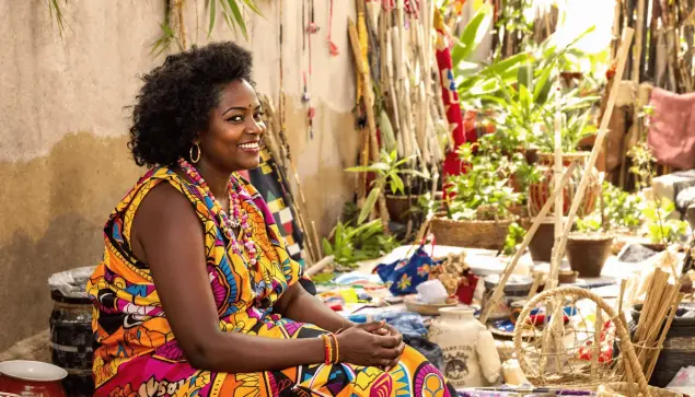 A woman in colorful traditional clothing sits peacefully outdoors among native plants and handmade crafts, bathed in warm sunlight.

