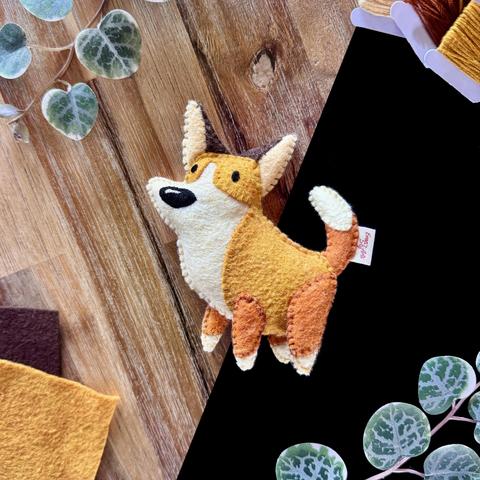 Picture of a handmade felt corgi plushie in front of a black and wooden background, surrounded with decorative plants and assorted felt and threads. 
The corgi has big brown ears, the inside of them is cream-coloured. 
The corgi has a soft orange back and an ivory muzzle and belly. Its feet and tip of its tail are the same colour. 
The orange on its legs and tail are a darken shade than the body. 

Two small beady eyes and a big shiny black nose are embroidered on its face. 