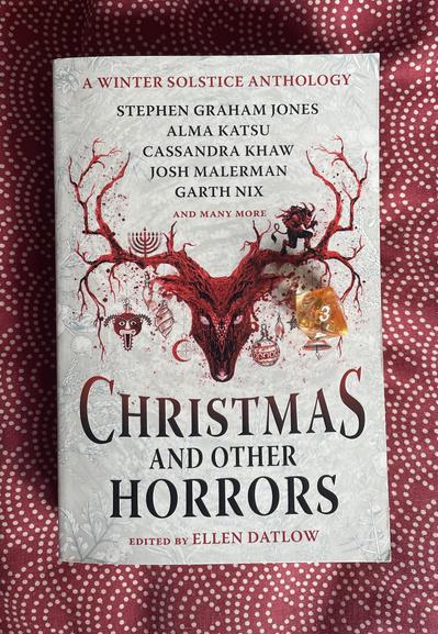 Book testing on a red and white patterned bedspread there is a yellow d8 sat on the cover showing the number 3. The cover is red and white and shows a horned animal head with winter holiday iconography hanging from it. 
A WINTER SOLSTICE ANTHOLOGY
STEPHEN GRAHAM JONES
ALMA KATSU
CASSANDRA KHAW
JOSH MALERMAN
GARTH NIX
AND MANY MORE
CHRISTMAS
AND OTHER
HORRORS
EDITED BY ELLEN DATLOW