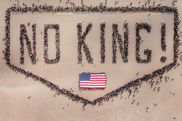 Thousands of people on a beach form the words "No King" with around a hundred people stretching out a big American flag. Arial view.