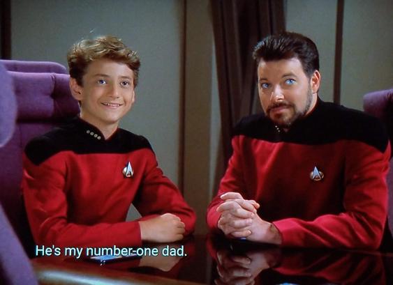 Star Trek The Next Generation scene, but with Hallmark channel vibes. We're "on the D" and "in the conference room" and Riker is shown sitting at the table, hands clasped, looking up with a muted smile. To his left, also sitting and w/ arms pleasantly on the table, is a smiling and happy looking young man. He's maybe 13 or 14 years old, and wearing the same red uniform and comm badge as Riker. Caption is centered over the little guy, and reads, "He's my number-one dad."