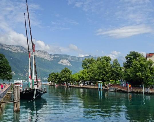 Am Kanal in Annecy. Links ist ein Segelboot im Hintergrund der See und die Alpen. 