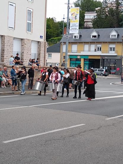 Une photographie d'une banda composée de six musiciens. Deux femmes de chaque côté de la ligne joue des grelots. Sur la même ligne deux autres femmes jouent un tambour profond et un homme de la caisse claire. En avant de la ligne, le meneur joue sur un tambour. Ils sont tous habillé en steampunk.