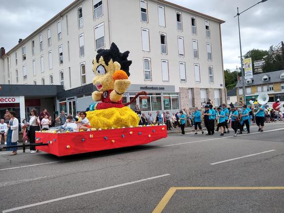Une photographie d'un char de carnaval représentant Son Goku, personnage de Dragon Ball Z, avec une queue de singe. Derrière, une banda habillée en t-shirt bleu et en pantalon noir joue de la musique.