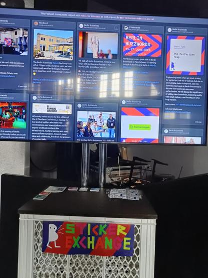 A photo of a display with the fediwall for Berlin Buzzwords on it. Underneath is a table with stickers on it and a colorful sign that says sticker exchange in letters made from tape.