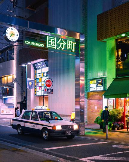 Nachtaufnahme aus dem Viertel Kokubuncho in Sendai, Japan. Ein beleuchtetes Schild mit Uhr zeigt den Viertelnamen. Im Vordergrund steht ein Taxi, rechts ein geöffneter Blumenladen mit Schaufenster und Kunde.