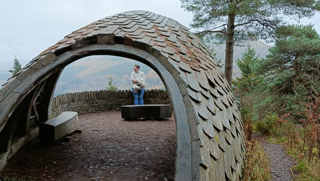Photo looking through a tunnel like structure in the Scottish countryside. In the distance is Éadaoín, who is leaning against a dry stone wall.