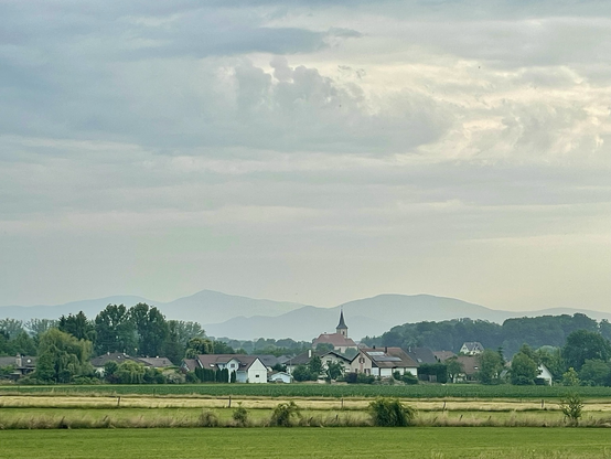 Lever du jour sur un village, église au centre et maisons autour. En fond de petites montagnes bleutées, ciel nuageux très au dessus
