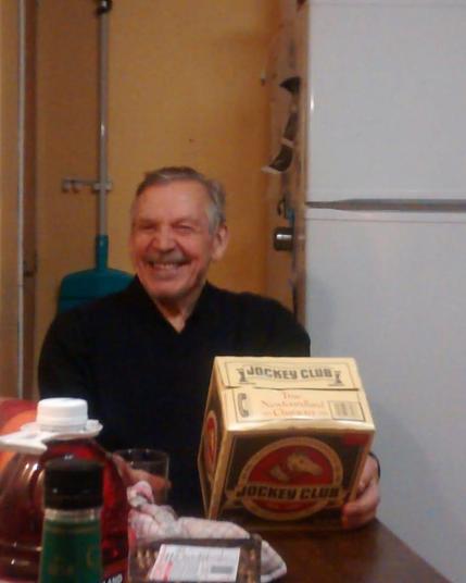 An older gentleman with short white hair and a black shirt sitting at a kitchen table with his hand on a box of Jockey Club beer, looking at the camera and laughing joyously.
