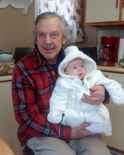 An older gentleman with longish grey hair and a red checked shirt sitting in a kitchen holding a baby in a white bunting bag.  He's clearly pleased as punch.  The baby would like to know what that over there is.

