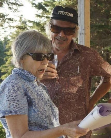 A man with grey hair and sunglasses and a maroon paisly short sleeved button up and base ball cap and a drink in his hand smiling at a woman with grey hair, sunglasses, and a blue patterned button up shirt singing while holding a song book.  They are standing on a patio on a nice sunny summer's day.
