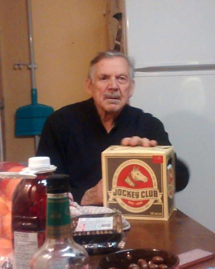 An older gentleman with short white hair and a black shirt sitting at a kitchen table with his hand on a box of Jockey Club beer.  He is looking into the distance and seems to be contemplating the horrors of the universe.
