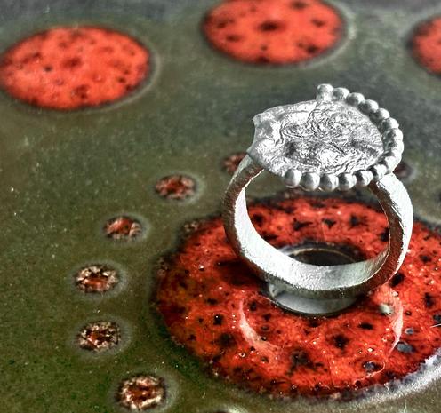 A silver ring featuring an ornate design sits atop a textured, dark surface with bright red circular patterns. The ring has a prominent decorative plate with a beaded edge.