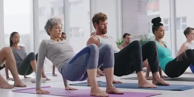 he image depicts a group of individuals participating in a yoga class. The setting is a bright, spacious room with large windows allowing natural light to fill the space. The participants are seated on yoga mats, performing a synchronized exercise. The person in the foreground is a woman with short gray hair, wearing a gray long-sleeve top and blue leggings, with her hands placed on the mat behind her, supporting her body in a backbend position. To her right, a man with a beard is wearing a whi…