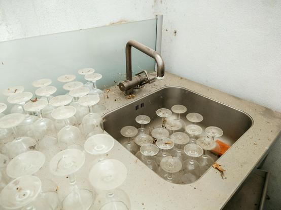 In der rechten Hälfte des Fotos eine ungepflegte Spüle, vollgestellt mit 16 auf dem Kopf stehenden Weingläsern. Links davon auf der Arbeitsfläche der Spüle viele weitere Gläser, auch auf dem Kopf stehend. Die Gläser sind alle gleicher Machart.