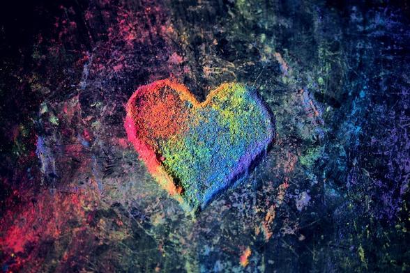 Image that shows a heart shape in rainbow colours. It's got a slightly raised edge and looks as if it is made of paint powder or chalk. There are splatters of the coloured powder around it.