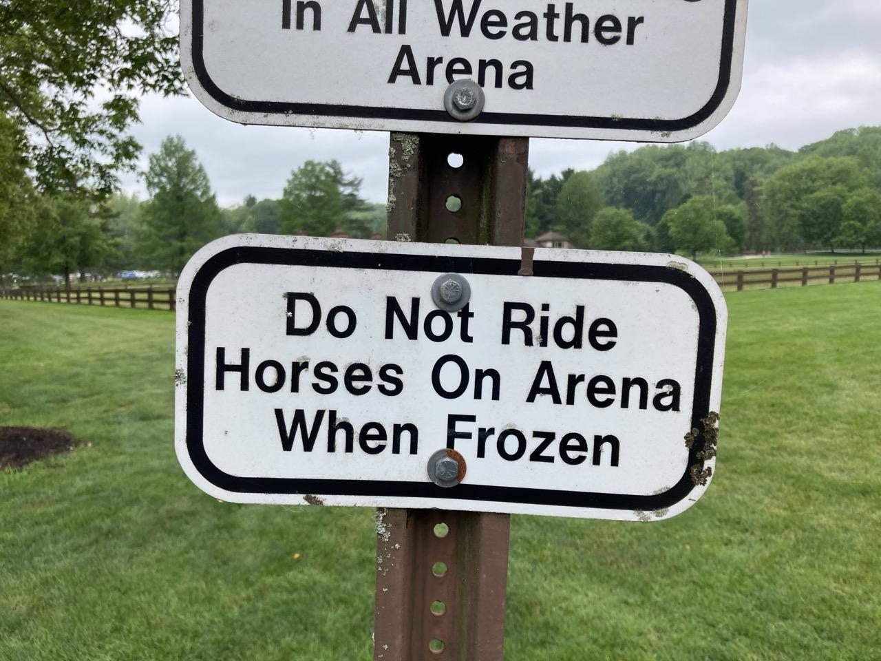 Signs on a post in front of an empty green field: In All Weather Arena Do Not Ride Horses On Arena When Frozen