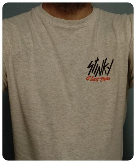 T-shirt blanc du groupe Stinky pour l'album "Of Lost Things". 
Le nom du groupe et de l'album sur le devant. 
Le logo du groupe en noir et celui de l'album en orange. 
Dans le dos c'est une illustration d'un loup sortant d'une porte avec des herbes sur les côtés. 
Toujours dans les tons orange/gris/noir. 
Ce n'est pas mon dessin préféré mais beau travail de l'artiste.