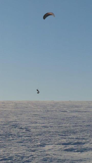 A snow kiter in mid air during a jump before a clear blue sky over crusty snow.
The high contrast and back light make him appear only as a dark silhouette and the kite is flying above in a black, orange and white design.