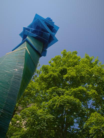 An upright photo from a very low perspective: a narrow “tree” of layered glass squares protrudes diagonally into the picture from the bottom left. The trunk consists of equal-sized squares of clear glass, which form a kind of helix as they become increasingly twisted. The crown is represented by larger squares of blue glass. Overall, however, it is a rather narrow and tall object. A large real tree can be seen behind and to the right of it, its structure, shape and color forming a beautiful contrast to the art object.

Ein Hochkant-Photo aus sehr niedriger Perspektive: diagonal von links unten ragt ein schmaler "Baum" aus geschichteten Glasquadraten ins Bild. Der Stamm besteht aus gleichgroßen Quadraten aus Klarglas, die durch zunehmende Verdrehung eine Art Helix bilden. Die Krone wird durch größere Quadrate aus blauem Glas dargestellt. Insgesamt ist es aber ein eher schmales und hohes Objekt. Dahinter und rechts davon ist ein großer echter Baum zu sehen und bildet durch seine Struktur, Form und Farbe einen schönen Konstrast zum Kunstobjekt.