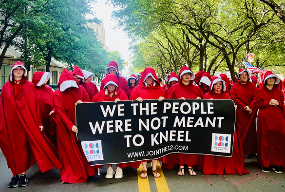 A row of women fill the image; they are wearing red cloaks and white bonnets of the Handmaid's Tale imagery. Fort Worth Texas. They carry a sign that says "We the people were not meant to kneel." www.jointhe12.com 