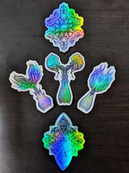 Photo of 5 holographic stickers laying on a dark wooden table. Top and bottom stickers are of mandalas. The middle 3 are 3 different mermaid tails.