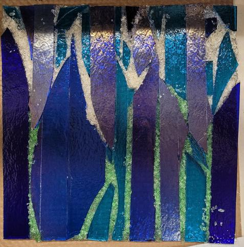 An unfired piece of glass art.  It is varying shades of blue shards up and down with space in between.  In the lower half, the spaces are filled with green frit (ground glass) and in the upper half, the spaces are filled with pale yellow frit.  Once it is fired, the light will "leak" through the frit