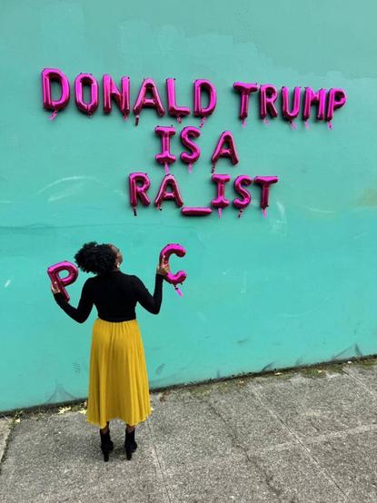 Eine schwarze Frau im gelben Rock  hält die Ballonbuchstaben "P" und "C" in den Händen, während sie vor einer Wand mit einem rosa Ballontext steht, auf dem steht: "DONALD TRUMP IST EIN RA_IST. Sie scheint sich nicht entscheiden zu können, welchen Buchstaben sie einsetzen soll.
Je nachdem, was sie einsetzt, hieße das fertige Wort dann entweder 
Rapist - Vergewaltiger oder
Racist  - Rassist