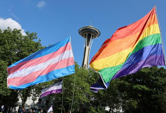 Three giant LGBTQIA+ Pride flags flying in front of the Space Needle. The Needle is in the center of the photo, framed by the flags in the foreground. A general rainbow Pride flag dances in the wind on the right side, closest to the camera. On the left, just a little behind the rainbow flag, a Trans Pride flag waves majestically just a little behind the rainbow flag. An Asexual Pride flag flutters beneath the Trans Pride flag, furthest back from the camera.