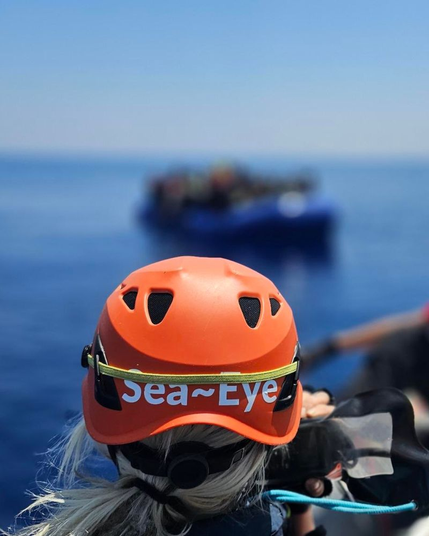 Von hinten ein*e Seenotretter*in mit einem orangen Schutzhelm, auf dem in weiß Sea~Eye steht. Die Person blickt in Richtunges blauen Schlauchboots voller Menschen, die nur schemenhaft zu erkennen sind, da dieser Bereich des Fotos entweder unscharf aufgenommen oder nachträglich geblurrt wurde.