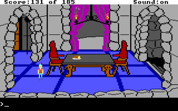 The dining hall of Dracula's castle in the published version of King's Quest 2. The walls of the room are light gray, and the dining table has a dark gray top and brown legs. Overhead is a candelabra with animated fire and smoke coming from the candles.