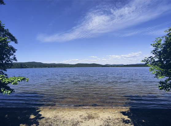 Foto desde la playa del Lac St-Charles enmarcada por árboles. Al fondo, cerros igualmente arbolados y el cielo azul en un día soleado y despejado. Oficialmente este es el km 32 del parque lineal.
