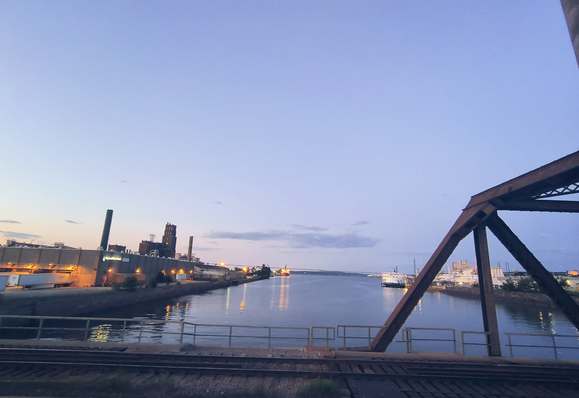 Embocadura del río San Lorenzo al anochecer. En ella hay al menos dos barcos y, a la izquierda, la fábrica de papel. La imagen está tomada desde el puente Samson, en Limoilou, con sus vías de tren. A unos pasos se encuentra el km 0 del parque.