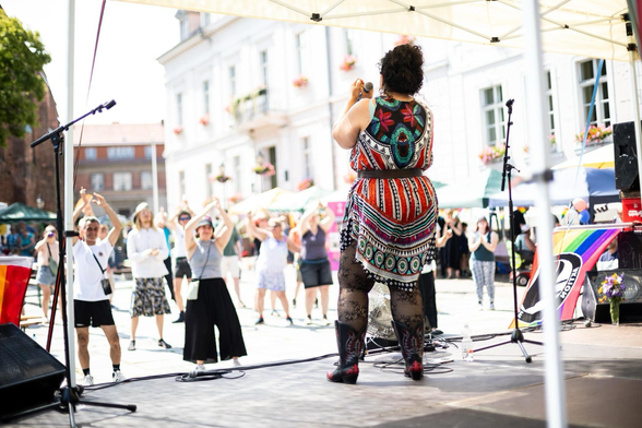 Die Sängerin Faravaz steht in buntem Kleid auf der Bühne, rechts eine Regenbogenfahne, vor ihr der Marktplatz von Bad Freienwalde und tanzendes Publikum. 