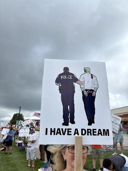 Foto.
Menschen demonstrieren. Eine Frau trägt ein Plakat mit der Aufschrift: I have a dream. Darüber sind zwei Personen von hinten gezeichnet: Ein Polizist führt Donald Trump in Handschellen ab.