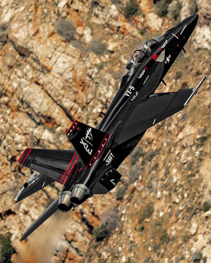 Texte alternatif (ALT) en français pour l’image :

Avion de chasse biplace noir, décoré de marquages blancs et rouges de l’US Navy, en vol ascendant à grande vitesse devant une paroi rocheuse. Les deux réacteurs laissent échapper une traînée de fumée, l’avion étant incliné sur l’aile gauche. Les pilotes sont visibles sous la verrière.