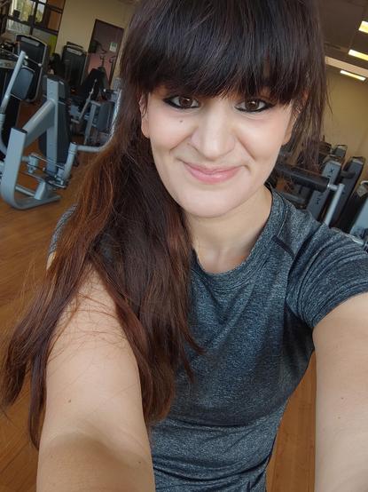 Selfie souriant pris dans une salle de sport. Une femme en tenue de sport grise, les cheveux longs attachés sur le côté, se tient devant plusieurs machines de musculation