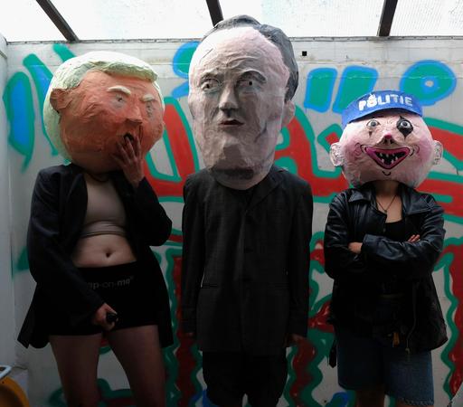 3 people with huge masks of mark rutte, donald trump and a cop, standing together against a wall. Donald trump is wearing a strap-on.