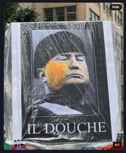 Photo of a board at a demo (no kings?) 

Picture on board has trump dressed & looking like Mussolini, but with bright orange cheeks with words below 

“IL DOUCHE”