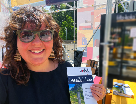 Selfie von Andrea Urthaler mit der aktuellen Ausgabe des bidok LeseZeichen in der Hand. Andrea lacht in die Kamera. Sie trägt eine Sonnenbrille. Im Hintergrund erahnt man die Bühnen-Konstruktion des Symposiums.