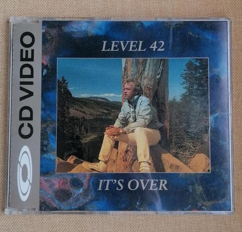 CD single for Level 52 "It's over" photo of bassists Mark King in casual wear sitting on a sandstone rock with a forest and mountains in the distance. Inset in blue painted art. Gold vertical flash on the left hand side proclaim "CD Video"