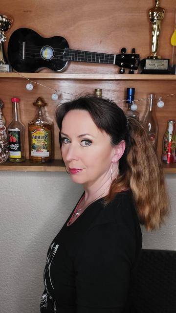 Martina steht im Partykeller. Hinter ihr im Regal eine Lichterkette, diverse Flaschen, ein Oskar und eine Ukulele