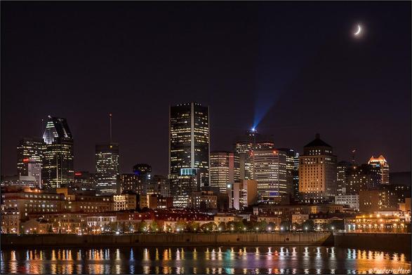 Night skyline of the city of Montreal with lots of lights of various colors in many tall buildings. Harbor in the foreground with lights reflecting of the water. A sliver of a moon visible in a corner.