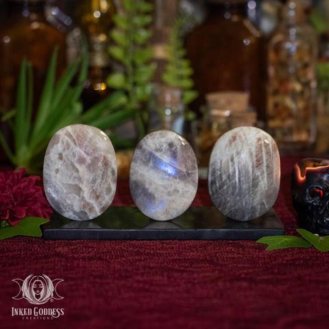 Three specimens of the "Rainbow Moonstone Gallet" product are arranged on a black stone slab. They are propped up to better catch the light and display their signature rainbow flash. They're in shades of white, grey, pink, and blue and polished to a mirror shine. Presented by Inked Goddess Creations.